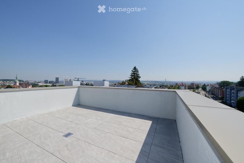 Flat rooftop terrace with light grey tiled flooring, white low walls, and a cityscape view under a clear blue sky.