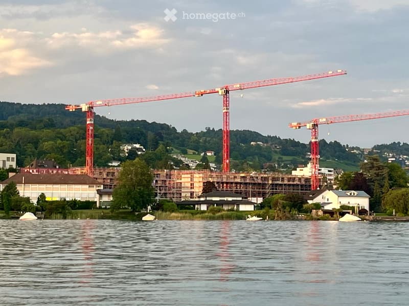 A construction site with two large cranes and scaffolding, situated near a body of water with surrounding greenery and existing buildings.