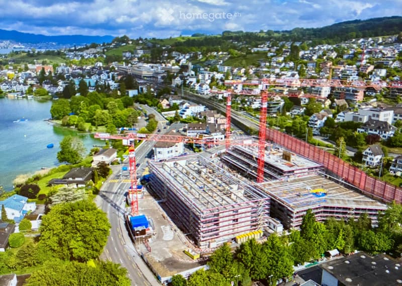 Aerial view of a city with a large construction site featuring cranes and scaffolding, surrounded by residential buildings, a lake, and green hills in the background.