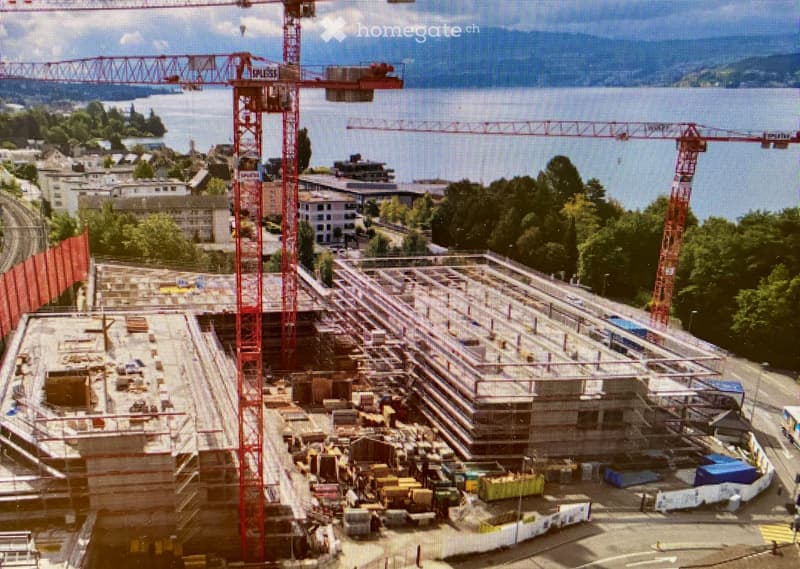 Aerial view of a construction site with cranes, scaffolding, and partially built structures, surrounded by greenery and a body of water in the background.