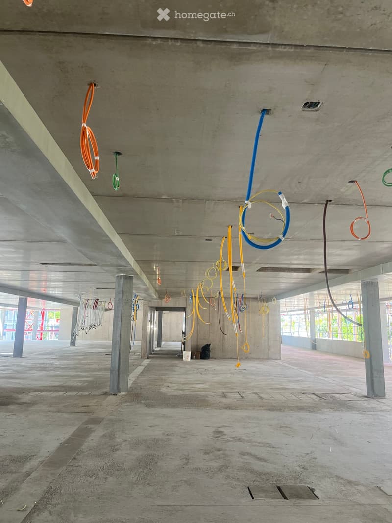 Empty concrete corridor with exposed electrical wires and cables hanging from the ceiling, supported by concrete pillars.