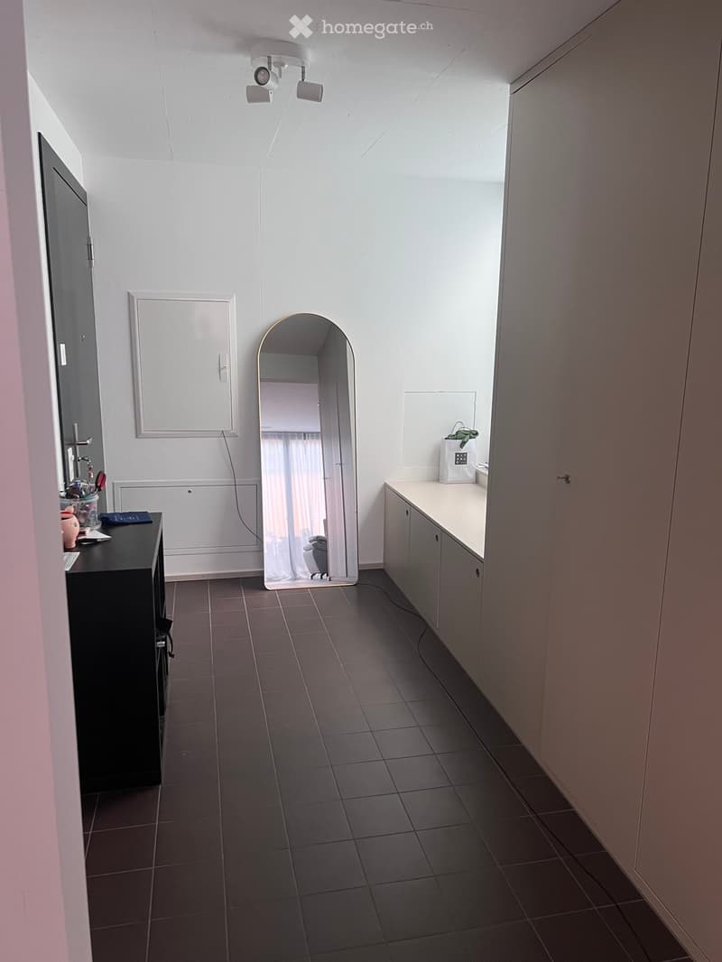 A modern hallway with dark tiled flooring, white walls, and built-in cabinets. Features include a large arched mirror, a small table with items, and a built-in cabinet with a countertop.