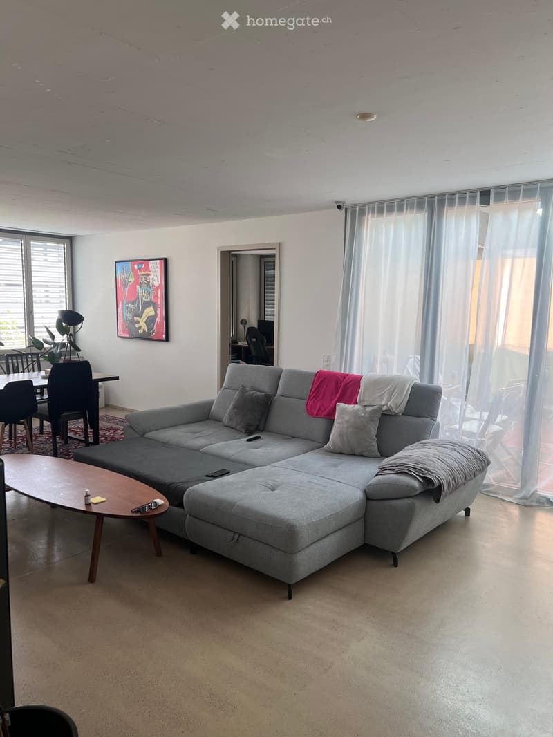 Modern living room with a grey sectional sofa, wooden coffee table, dining area with chairs, and large windows with sheer curtains.