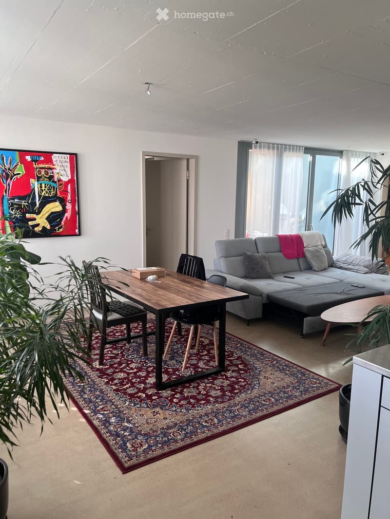 Modern living room with a grey sectional sofa, wooden coffee table, dining table with chairs, large area rug, and abstract painting on the wall.