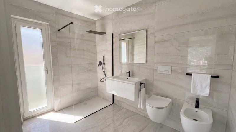 Modern bathroom with marble tiles, a glass-enclosed shower, a white sink, a toilet, a bidet, and a mirror above the sink. Towels are neatly placed on a rack.