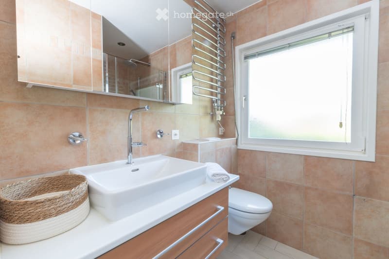 Modern bathroom with a large sink, chrome faucet, toilet, mirrored cabinet, and a wicker basket on the counter. The walls are tiled in warm beige tones, and there is a window with frosted glass for natural light.