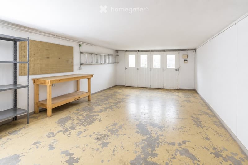 Empty room with white walls, a wooden table, metal shelving unit, and a large garage door with windows.