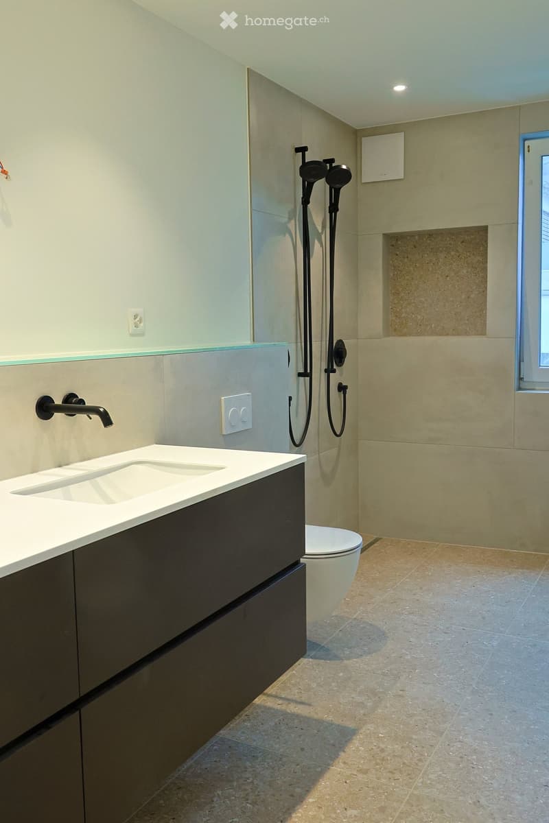 Modern bathroom with a white sink, black faucet, toilet, and a multi-head shower system. Light-colored tiles and a sleek design.