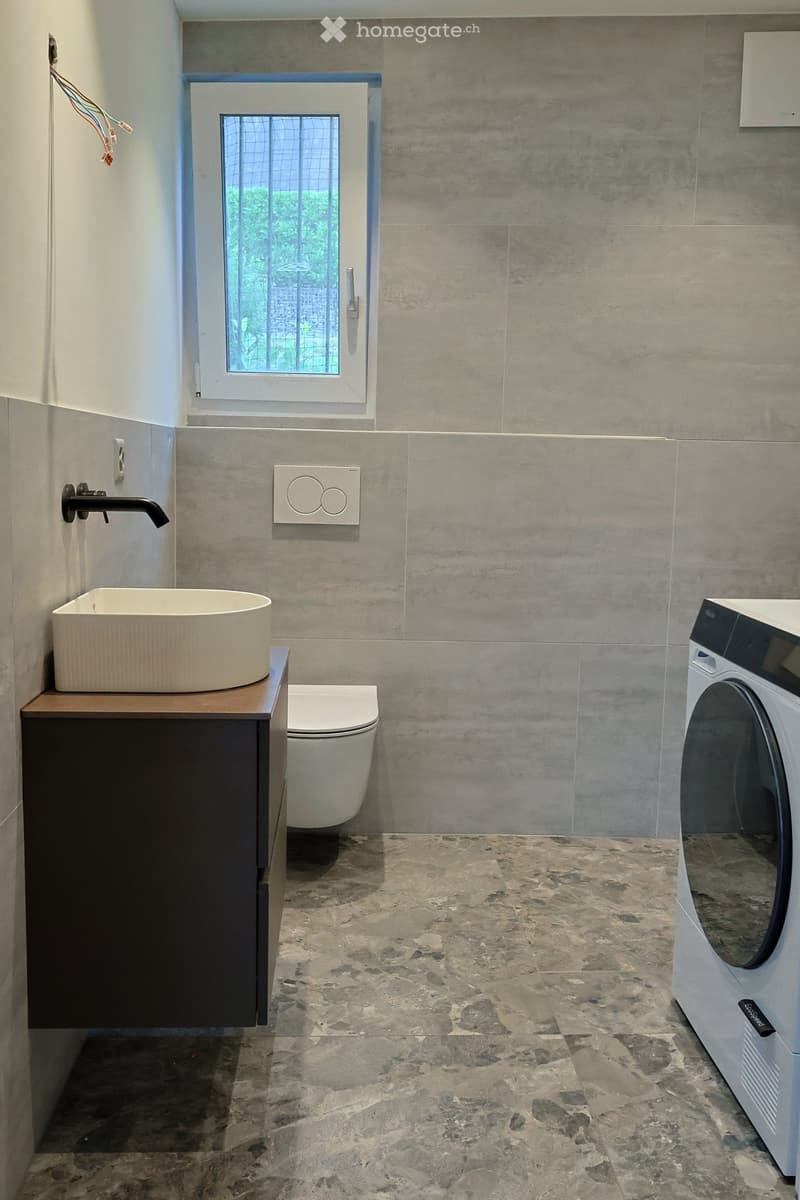 Modern bathroom with a white sink, black faucet, toilet, and washing machine. Features a window with a grid pattern and grey tiled walls and floor.