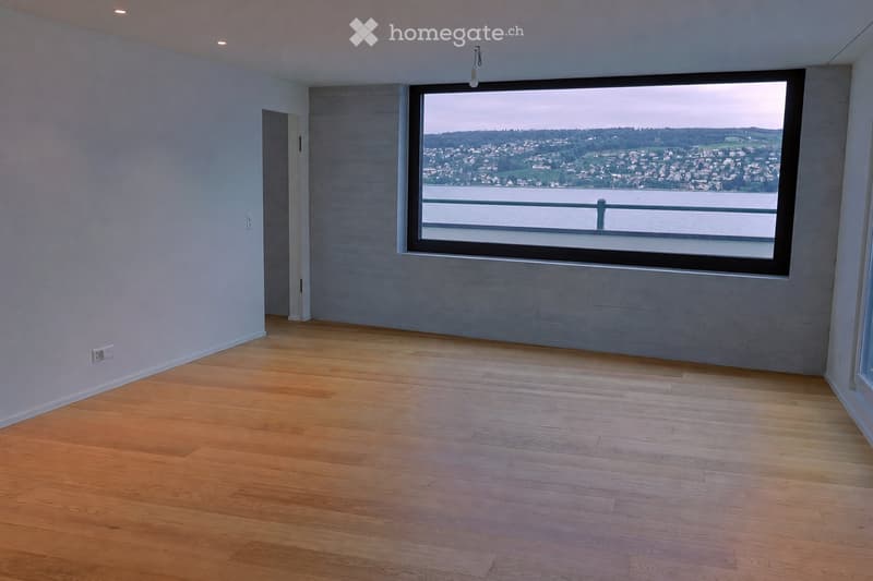 Empty room with wooden flooring, large window offering a scenic view of a city and water, and modern lighting fixtures.