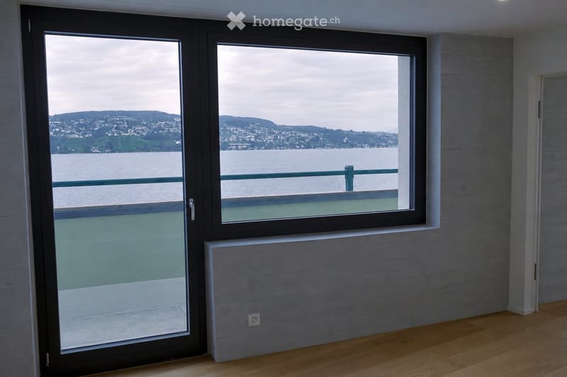 Empty room with wooden flooring, grey walls, large sliding glass doors leading to a balcony with a scenic view of water and distant hills.