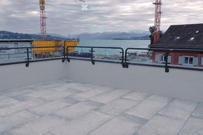 Roof terrace with a smooth concrete floor, metal safety railing, and a scenic view of a body of water and distant mountains.