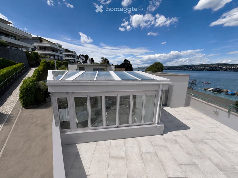 A modern glass-enclosed terrace with a retractable roof, overlooking a waterfront with boats and a distant view of the lake and hills.