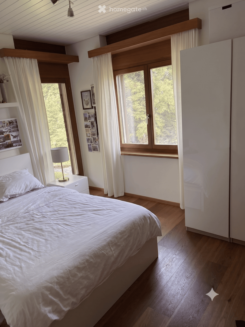 Modern bedroom with a large bed, nightstand, lamp, and large windows with curtains. The room features wooden flooring and white walls.