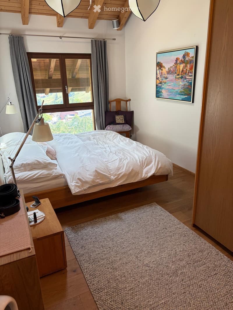 Cozy bedroom with wooden floors, a large bed with white bedding, a nightstand with a lamp, a small wooden chair with a cushion, a painting on the wall, and a gray rug on the floor.