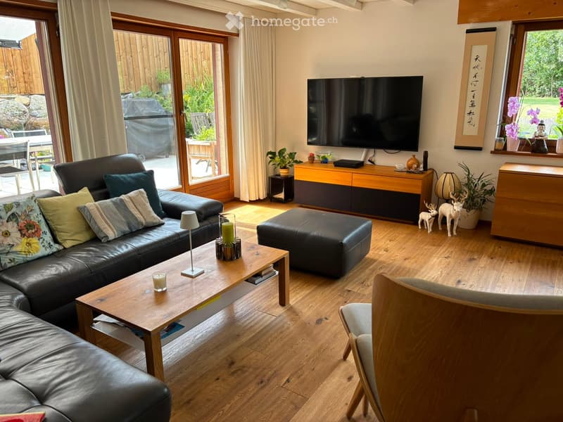 Modern living room with hardwood flooring, large windows, and a balcony door leading to a garden. Features a leather sofa, coffee table, ottoman, armchair, TV, sideboard, and decorative items.