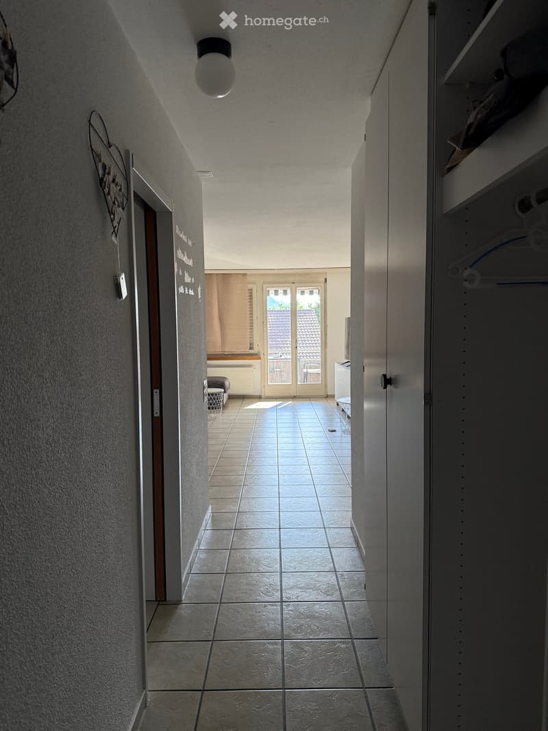 A narrow hallway with tiled flooring, white walls, and a ceiling light fixture. A closet with hanging clothes is visible on the right, and a door leads to another room at the end.