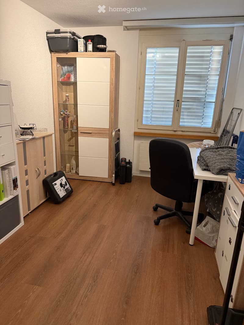 A well-lit room with wooden flooring, a desk with a chair, a cabinet, and a window with blinds.