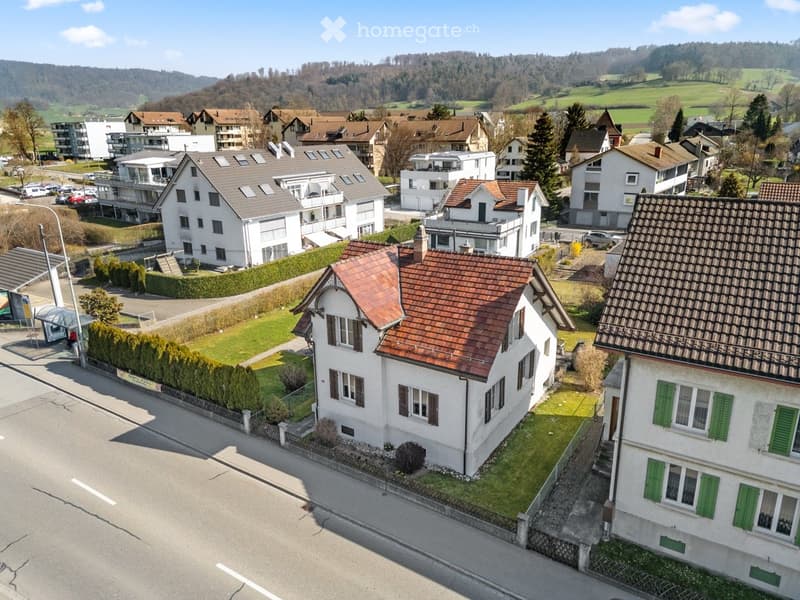 Aerial view of a residential area with multiple houses, green lawns, and a hilly landscape in the background.