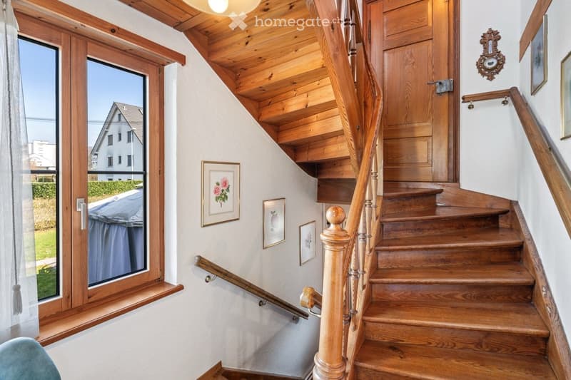 Wooden staircase with handrail and balusters, white walls, framed artwork, large window with view of the outside, and a light fixture on the ceiling.