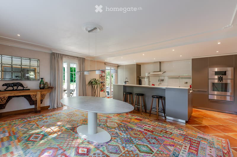 Open-plan living and dining area with a modern kitchen, central island, bar stools, round dining table, decorative vases, and a large mirror on the wall.