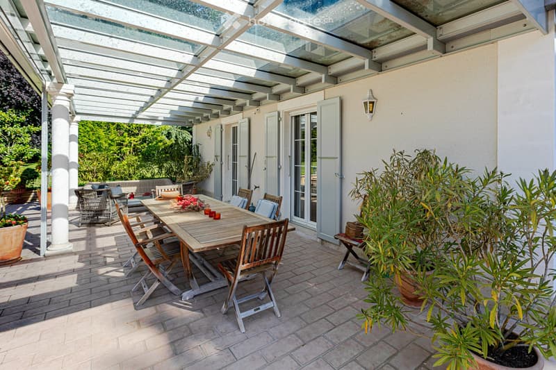 A spacious outdoor patio with a wooden dining table and chairs, surrounded by potted plants and a covered pergola. The area features a tiled floor and large glass doors leading to an indoor space.