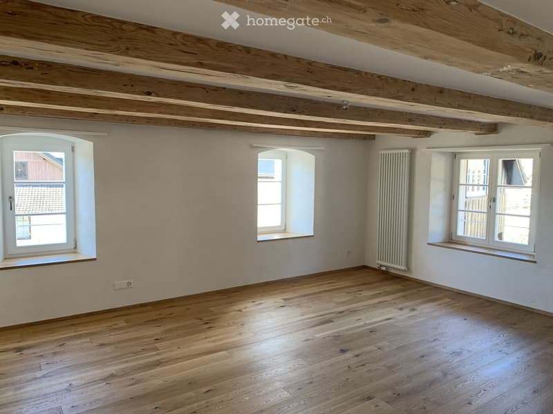 Empty room with wooden floor, exposed wooden ceiling beams, and several windows with white frames. The walls are painted in a light neutral color, and there is a radiator on one wall.