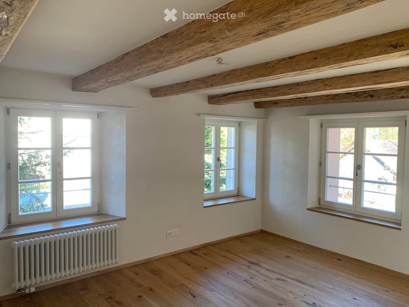 Empty room with wooden floor, exposed wooden beams on the ceiling, three windows with white frames, and a radiator on the left side.