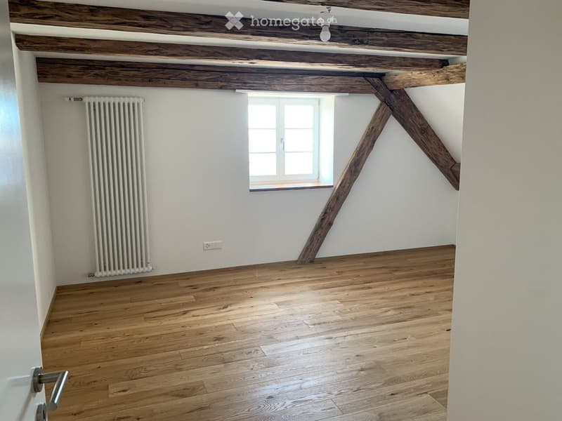 Empty room with wooden floor, white walls, exposed wooden beams, radiator, and a window with white frame.