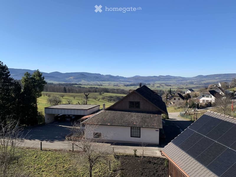 A two-story house with a garage, surrounded by a rural landscape with trees and distant hills.