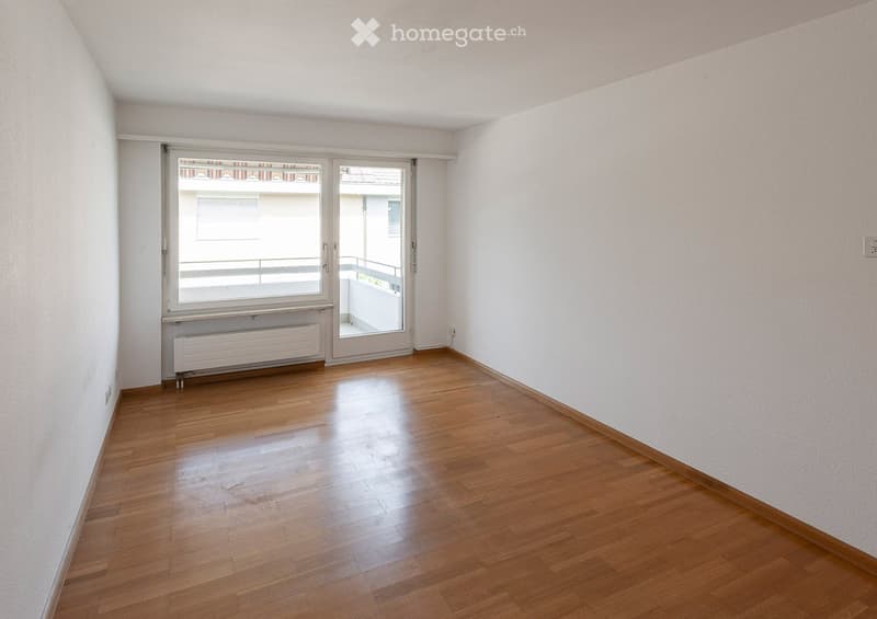 Empty room with wooden flooring, white walls, large window, and sliding glass door leading to a balcony.