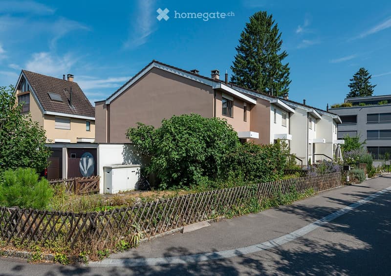 Residential street with multiple two-story houses, surrounded by greenery and a wooden fence.