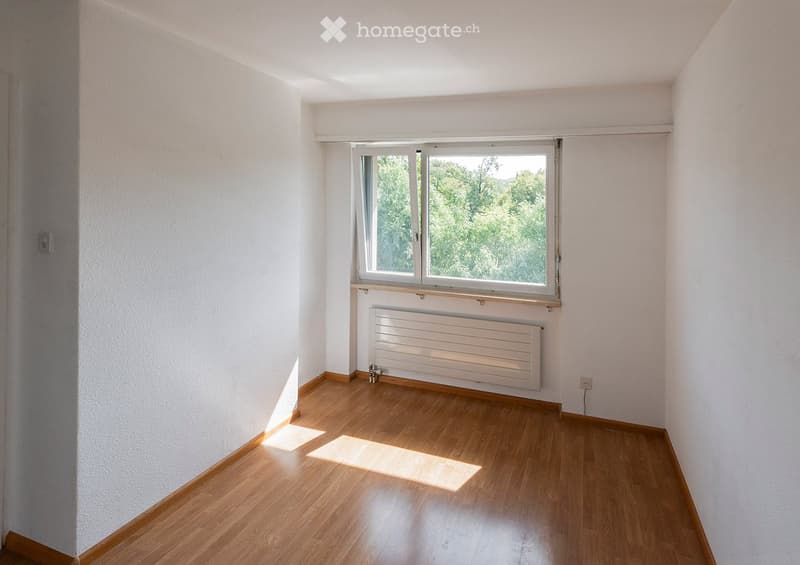 Empty room with light wooden flooring, white walls, a large window with a view of greenery, and a radiator below the window.