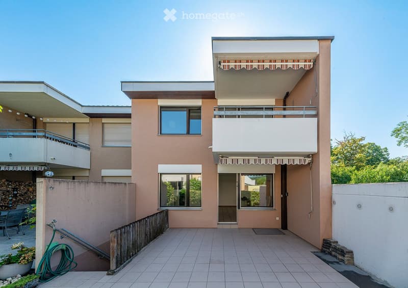 Two-story house with beige facade, large windows, balconies, and an awning on the upper level. The ground floor features a covered entrance and a patio area with outdoor furniture.