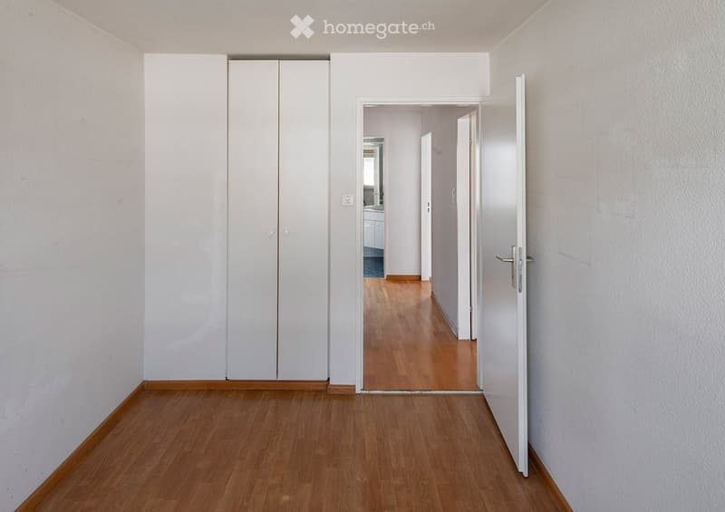 Empty hallway with wooden flooring, white walls, and a closed white closet on the left. An open door leads to another room with visible wooden flooring and white walls.