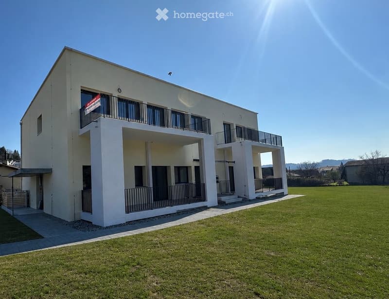 Modern two-story building with white exterior, multiple balconies, and a spacious lawn in front.