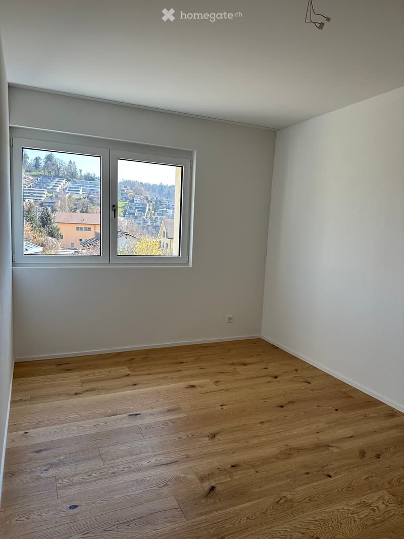 Empty room with wooden flooring, white walls, and large windows offering a view of a residential area.