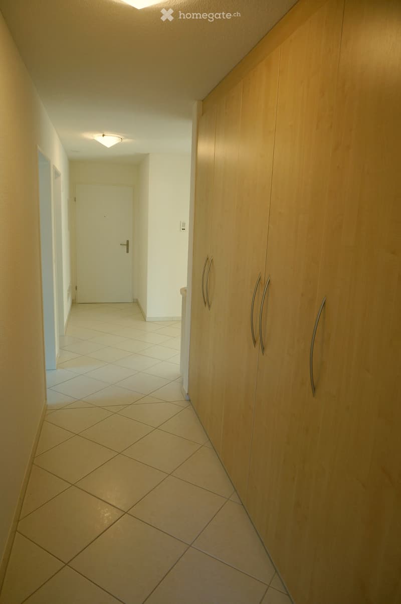 A clean, well-lit hallway with tiled flooring, light-colored walls, and wooden built-in storage units with silver handles. A closed door is visible at the end of the hallway.
