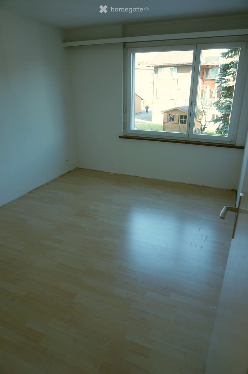 Empty room with light wooden flooring and white walls, featuring a large window with a view of the outside area.
