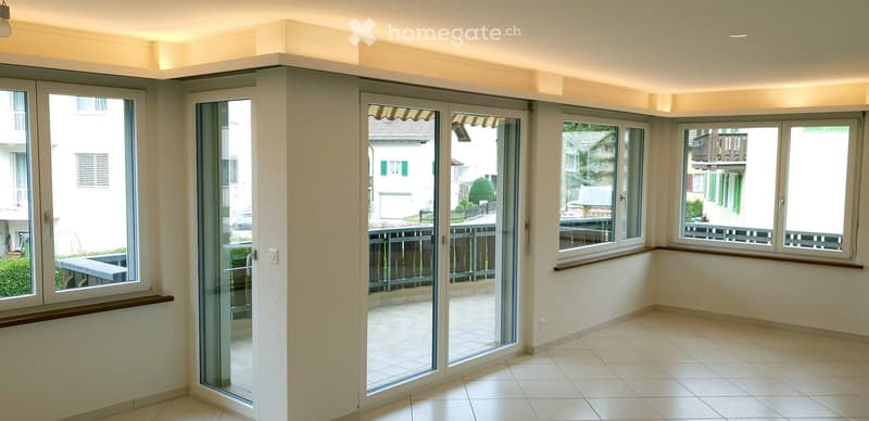 Empty room with large windows and sliding glass doors leading to a balcony. Light-colored tiled floor and white walls.