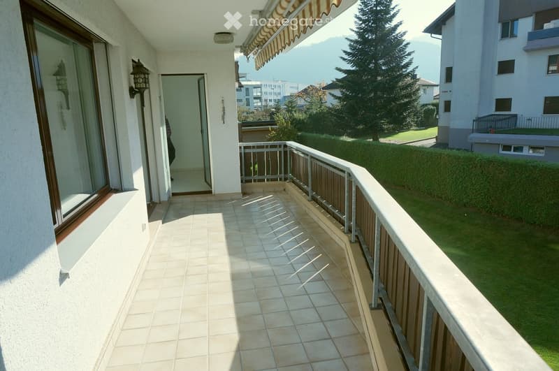Balcony with tiled floor, metal railing, and striped awning, overlooking a green lawn and neighboring building.