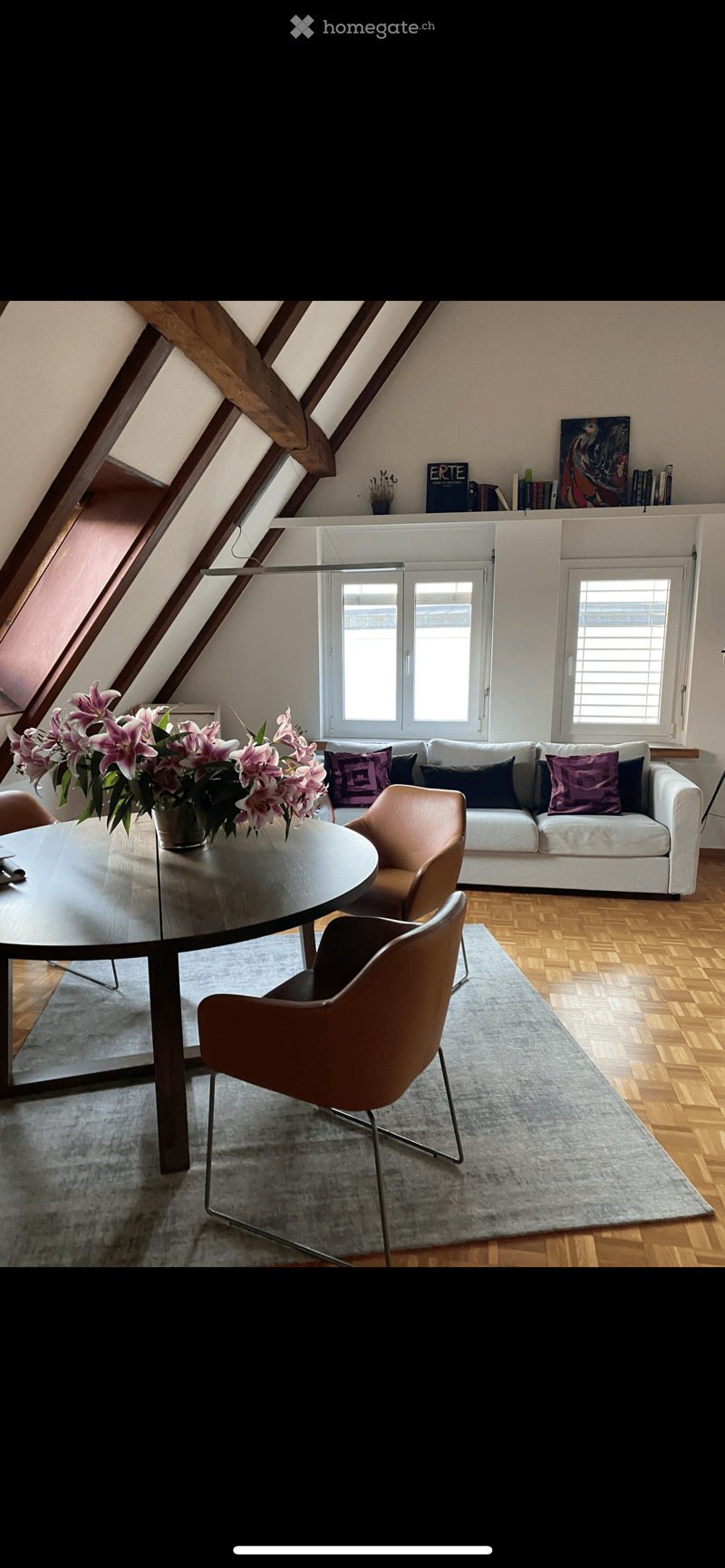 Modern living room with wooden floors, large windows, a white sofa with colorful cushions, a round glass table, and a vase with pink flowers.