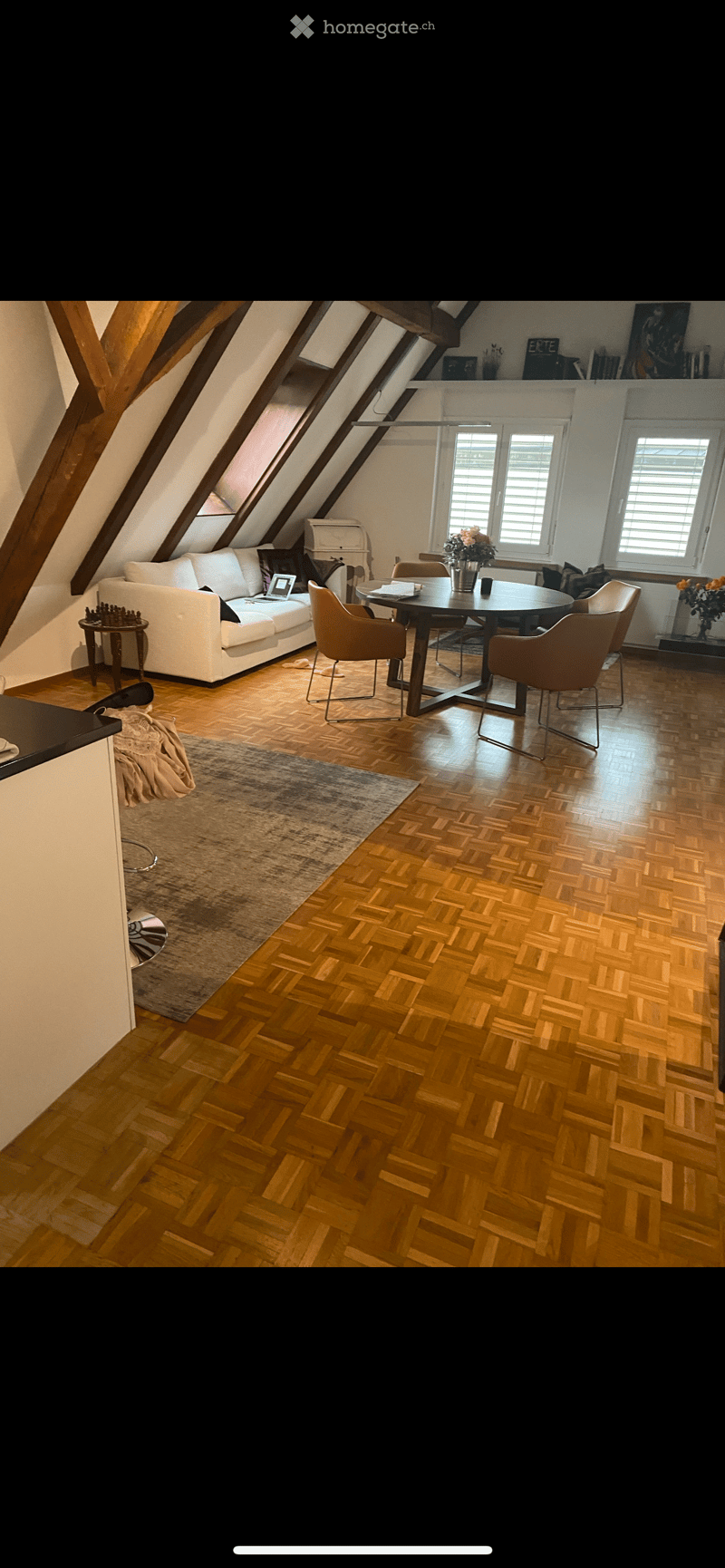 Spacious attic room with wooden herringbone flooring, white sofa, round dining table with four chairs, laptop on sofa, vase with flowers, small side table, and a large area rug.