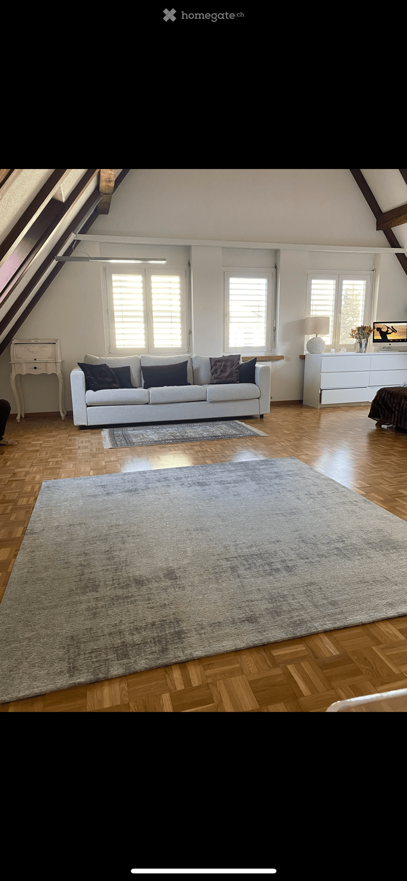 Bright living room with wooden flooring, large windows, a white sofa with multiple cushions, a coffee table, a large area rug, a side table with a lamp, and a television on a stand.