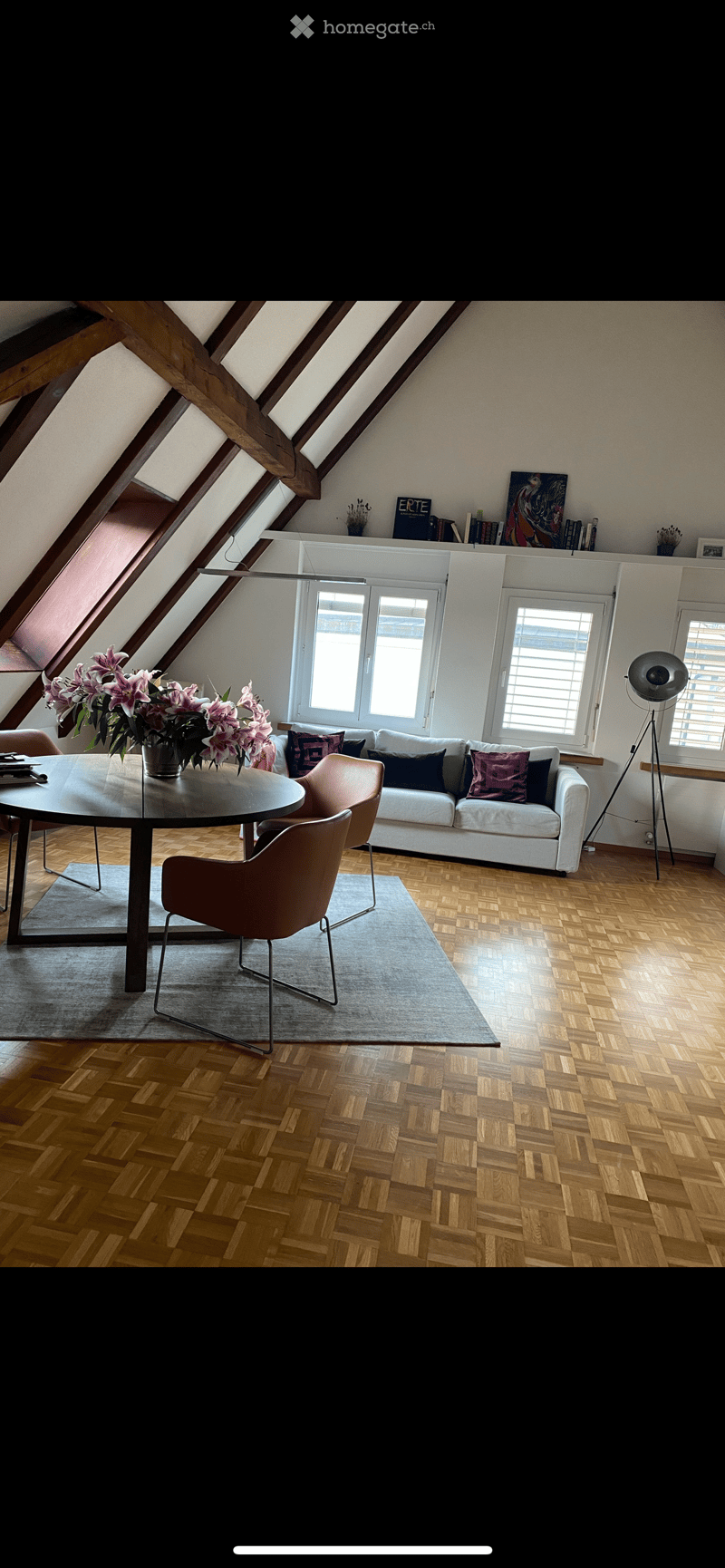 Modern living room with wooden herringbone flooring, white sofa with colorful cushions, round glass table with chairs, and a built-in bookshelf with books and decorative items.