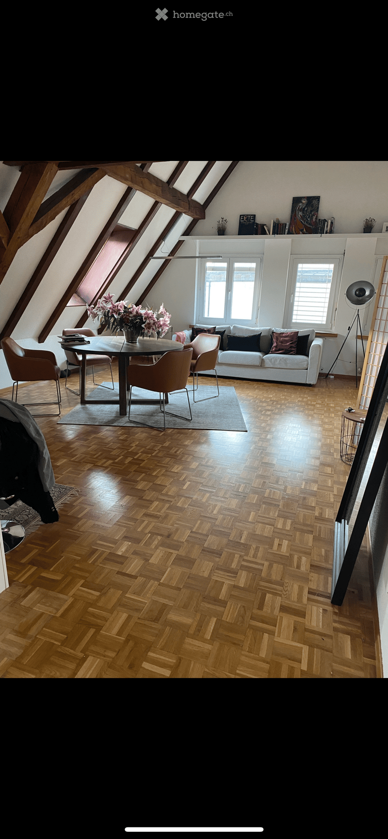 Bright living room with wooden herringbone flooring, white walls, and large windows. Features a white sofa with colorful cushions, a round dining table with four chairs, and a vase of pink flowers on the table. A floor lamp and bookshelf are also visible.