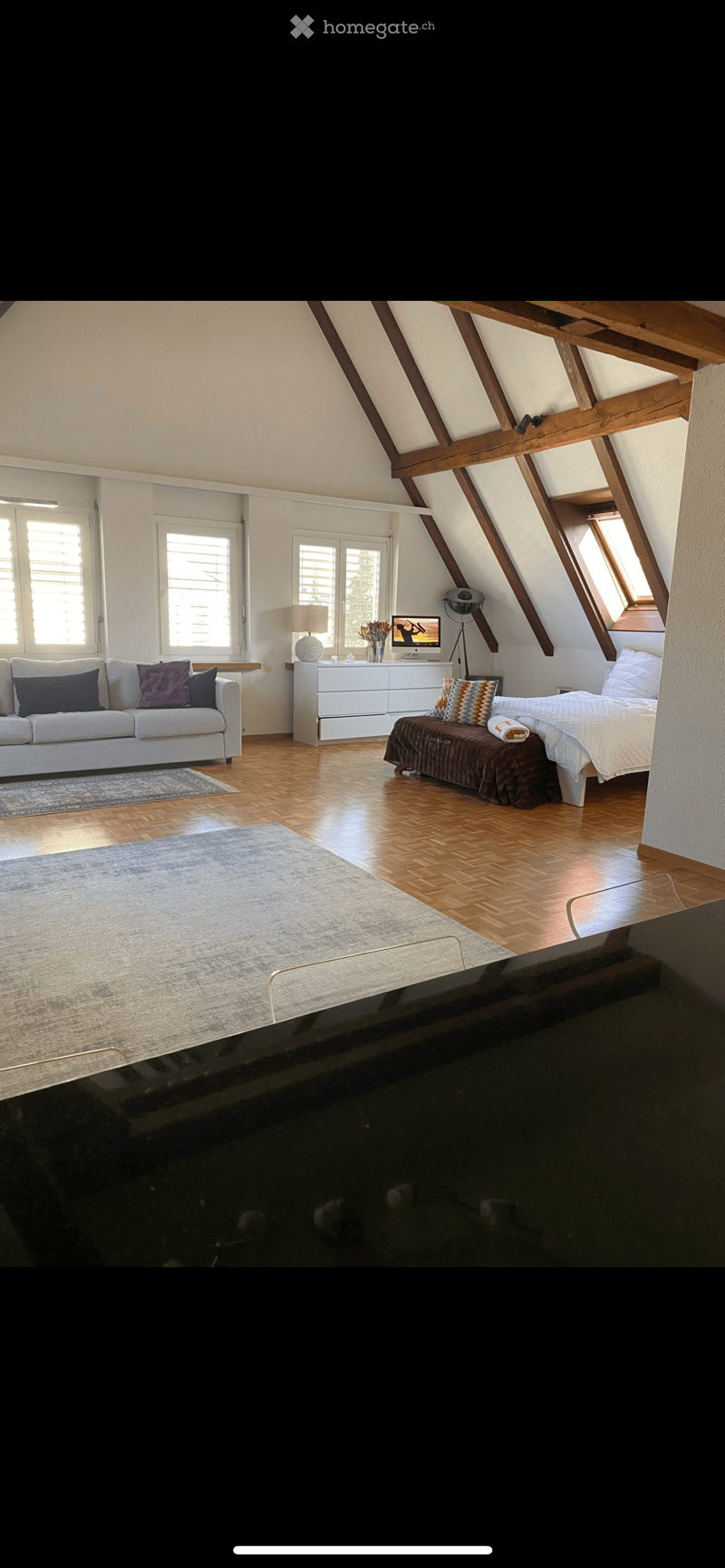 Spacious attic room with wooden floors, slanted ceilings, large windows, a sofa, a bed, a dresser, and a computer monitor.