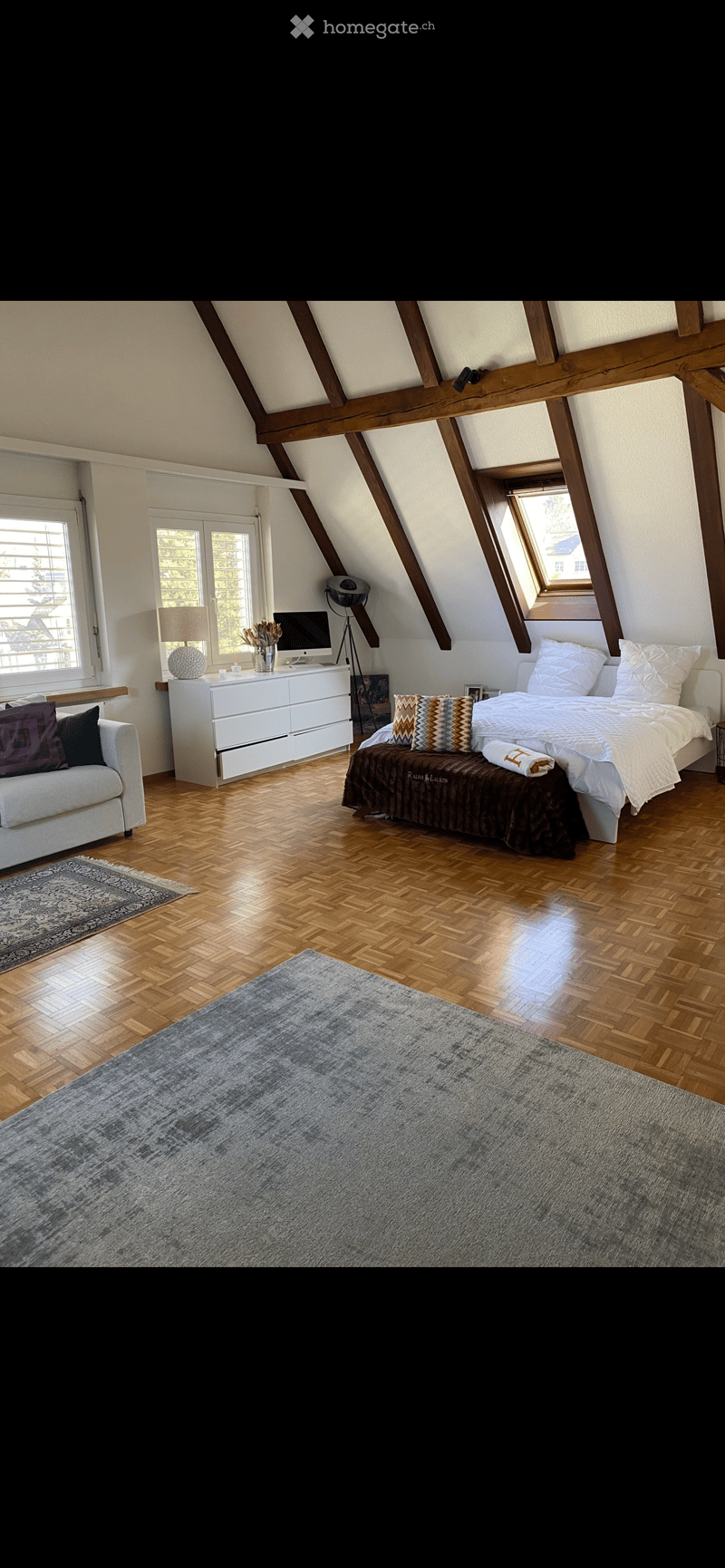 Bright attic bedroom with wooden slanted ceilings, large windows, a double bed, a sofa, a dresser, and a computer on a small table.
