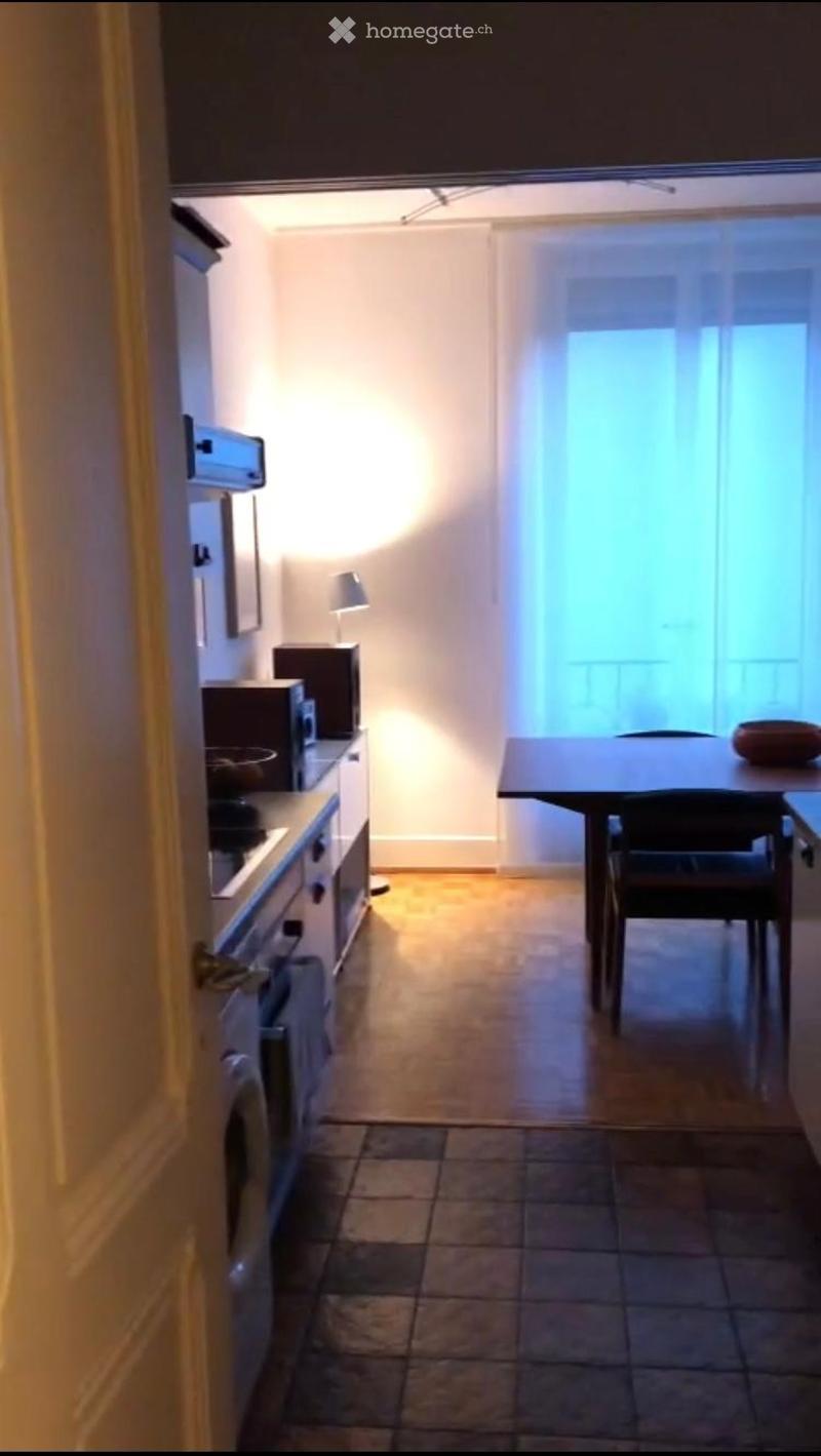 A small, modern kitchen with a table and chair, microwave, oven, fridge, and a lamp on the counter.