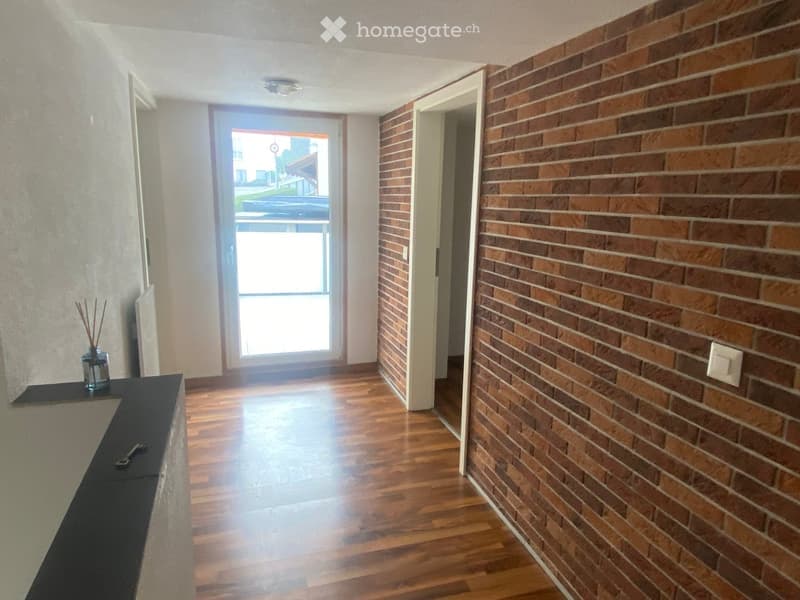 Hallway with wooden flooring, brick wall on one side, and a door leading outside. A small shelf with decorative items is visible on the left.